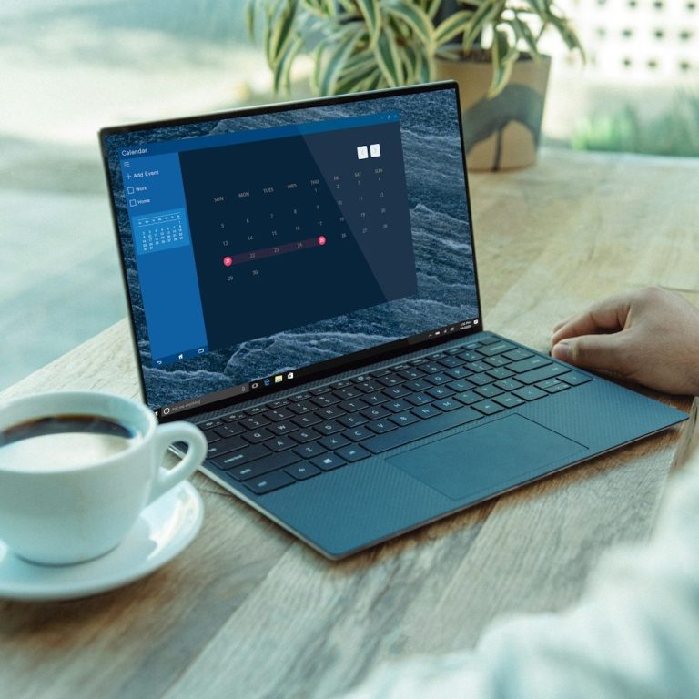 Desk with a cup and saucer, next to an open dark grey laptop with the screen on. Plant in background.